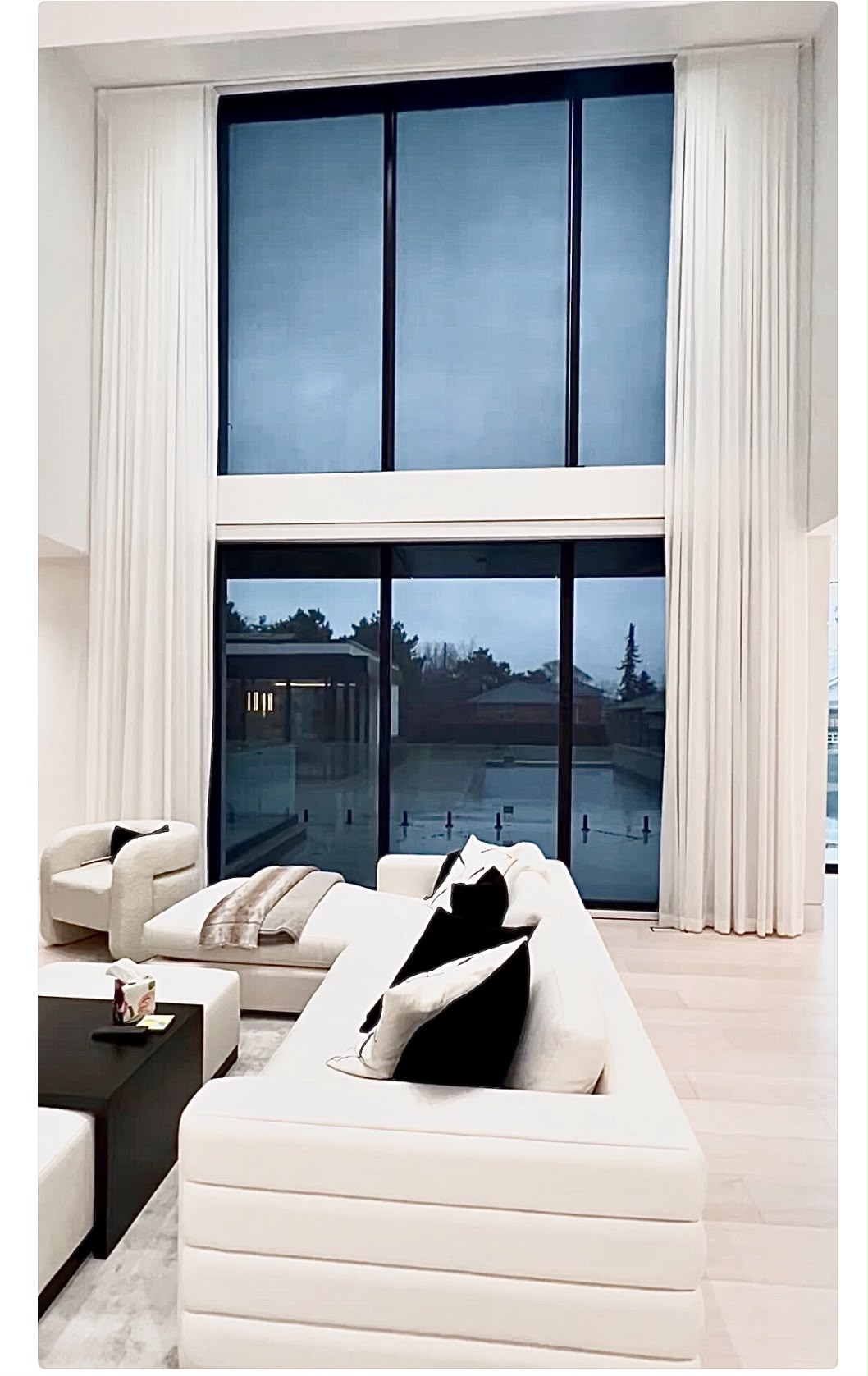 Roller shades and ivory drapery framing double-height windows in a contemporary living room